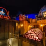 Une scène nocturne animée à Disneyland Paris avec l'enseigne lumineuse « Gusteau's » au-dessus d'une façade de style français, avec deux visiteurs levant les yeux avec admiration.