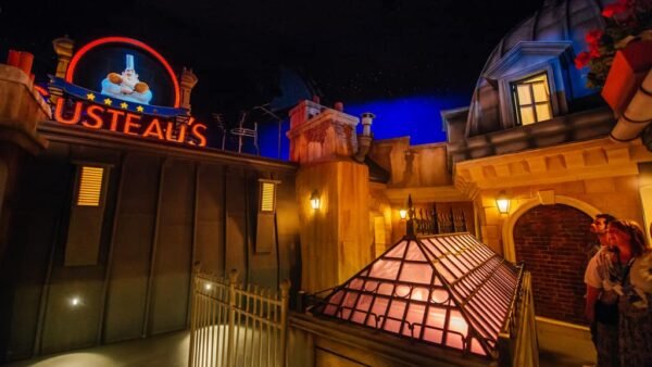 Une scène nocturne animée à Disneyland Paris avec l'enseigne lumineuse « Gusteau's » au-dessus d'une façade de style français, avec deux visiteurs levant les yeux avec admiration.
