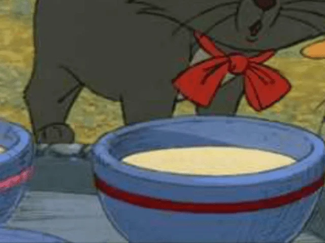 Un chat gris animé portant un nœud papillon rouge s'accroupit entre des bols de lait, semblant prudent ou sournois dans cette scène d'un film Disney.