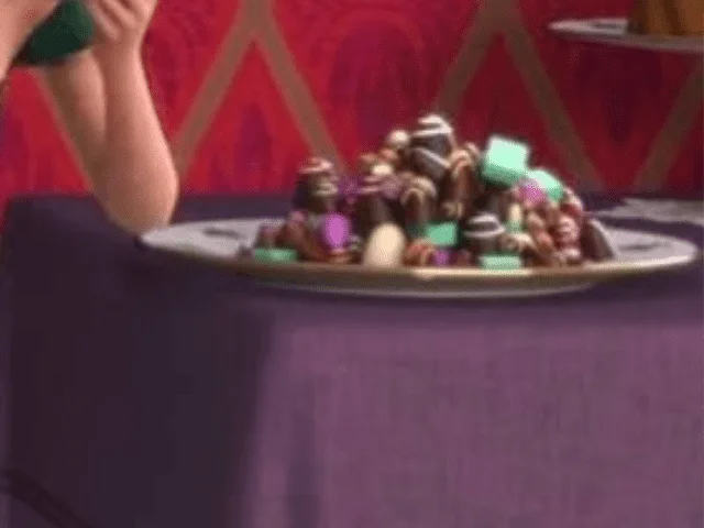 Un plateau rempli d'une variété de chocolats et de bonbons colorés inspirés des films Disney, partiellement masqué par le bras d'une personne au premier plan sur un fond aux motifs vibrants.