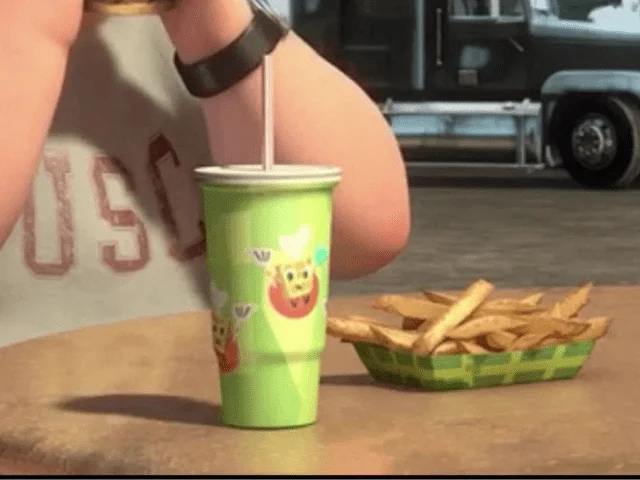 Gros plan d'une main tenant une paille placée dans une tasse verte avec des autocollants de fruits sur le thème des films Disney, à côté d'un panier de frites sur une table, avec un véhicule flou en arrière-plan.