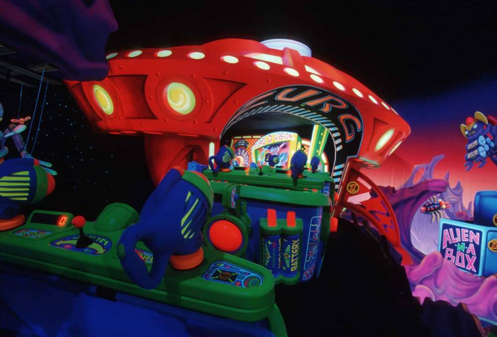 Manège animé dans un parc à thème à Disneyland Paris avec une entrée de vaisseau spatial, ornée de motifs extraterrestres colorés, menant à un tunnel avec des jeux de tir de style arcade. Les visiteurs visent des cibles depuis des gamepods futuristes