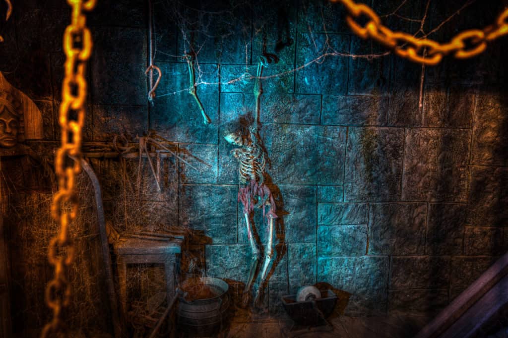Une scène de donjon effrayant à Disneyland Paris avec un squelette enchaîné à un mur éclairé par une faible lumière, entouré de murs en pierre et de chaînes suspendues, créant une atmosphère étrange.