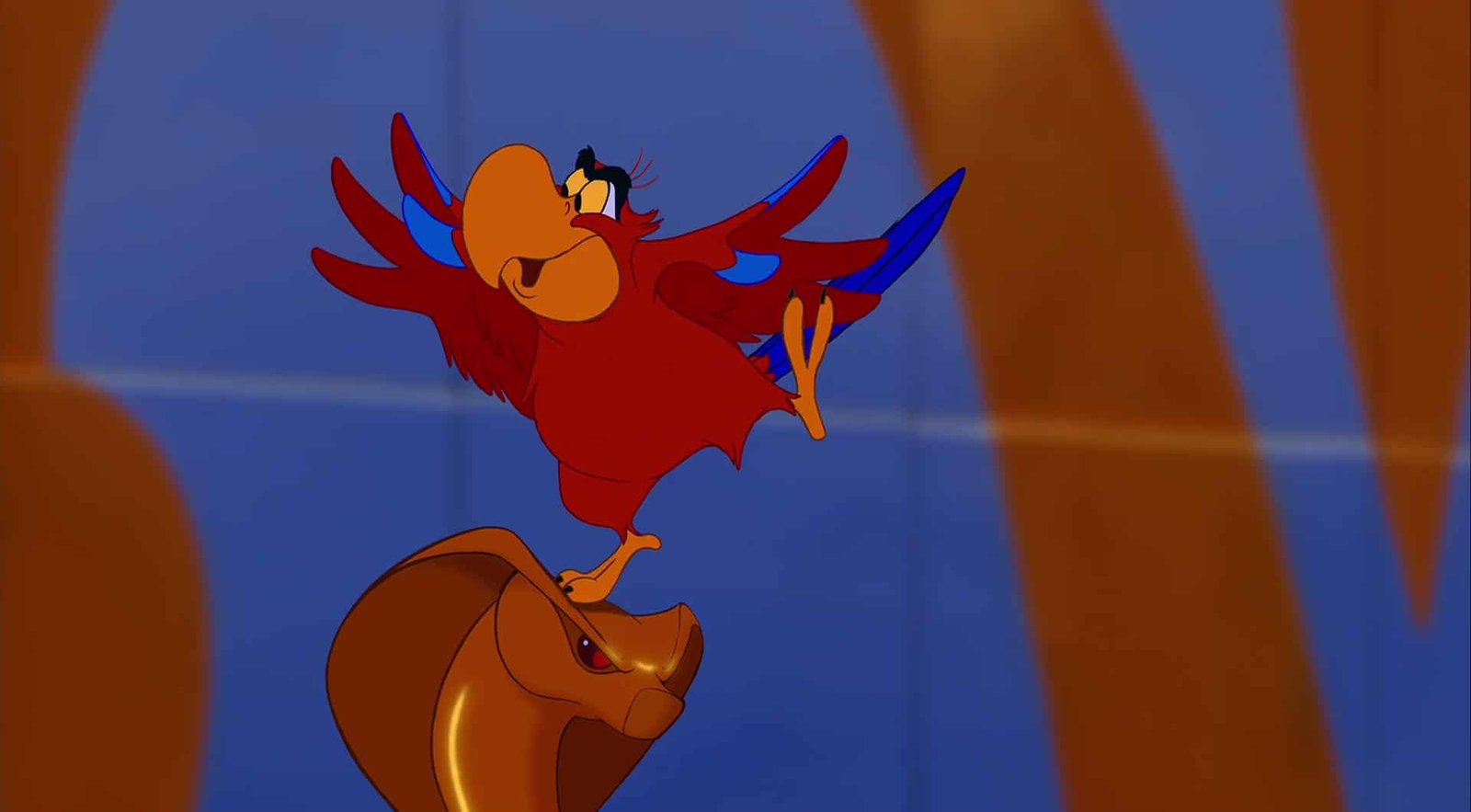 Iago, un perroquet rouge, vole énergiquement avec les ailes déployées, tandis qu'Abu, un petit singe brun, l'observe attentivement d'en bas, dans un décor désertique depuis un
