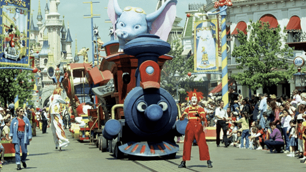 Un défilé coloré à Disneyland mettant en vedette un grand char de cirque toon avec un éléphant bleu ressemblant à Dumbo, entouré d'artistes aux costumes éclatants, et des spectateurs excités bordant la rue.