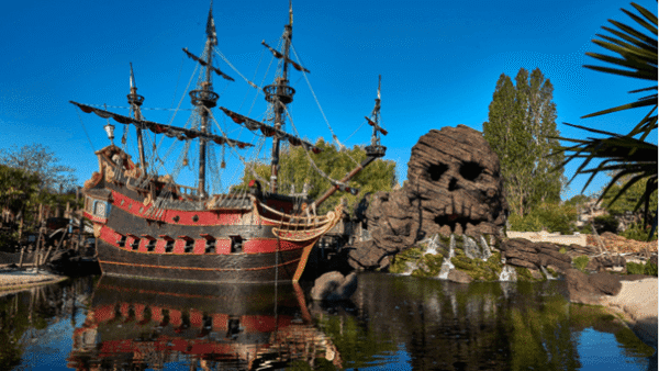 Un grand bateau pirate orné ancré dans une eau bleu clair, avec des mâts et des cordes détaillés, près d'un rivage rocheux avec une verdure luxuriante et une grande formation rocheuse en forme de crâne.