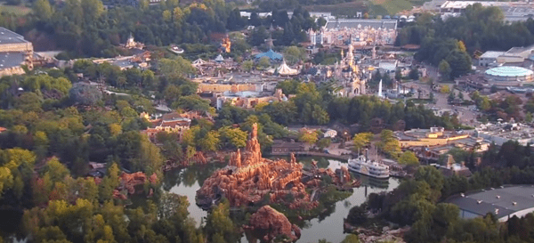 Vue aérienne d'un parc à thème révélant des secrets comme un château, une grande structure de montagne, des plans d'eau et diverses attractions nichées au milieu d'arbres verdoyants.