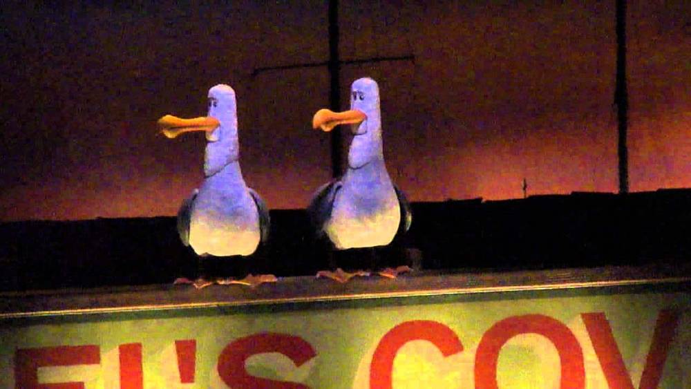 Deux personnages de mouettes animés perchés sur un panneau indiquant « la crique » à Disneyland Paris, provenant peut-être d'une production théâtrale ou cinématographique, éclairés sous l'éclairage de la scène.