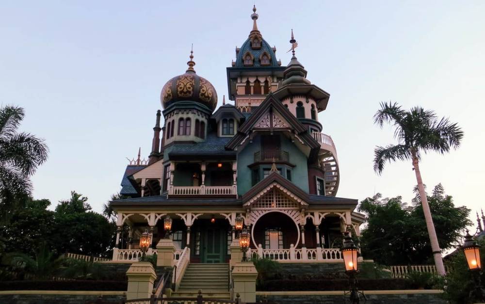 Un manoir victorien orné au crépuscule, présentant des détails architecturaux complexes, de multiples tourelles et des palmiers au premier plan, rappelant le Manoir Fantôme de Disneyland Paris.