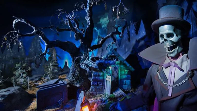 Une scène effrayante de Phantom Manor avec un squelette portant un haut-de-forme et un costume, entouré d'arbres étranges, de pierres tombales et d'un fond bleu brumeux.