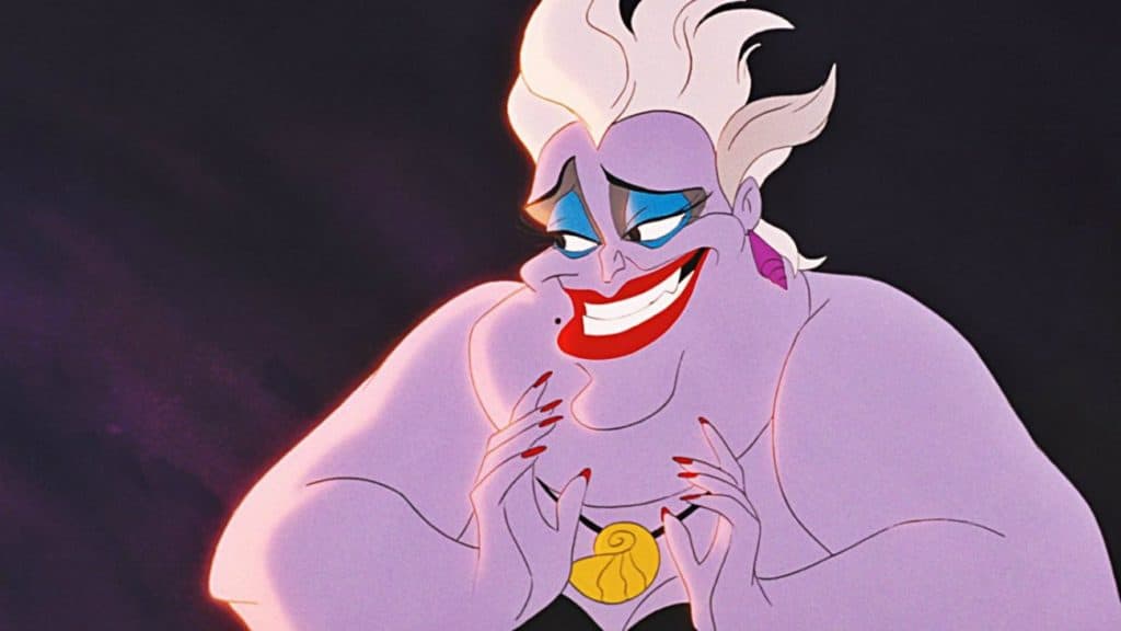 Ursula, une sorcière des mers du dessin animé Disney aux cheveux blancs et à la peau violette, sourit malicieusement tout en serrant un pendentif en forme de coquille de nautile doré, sur un fond sombre.