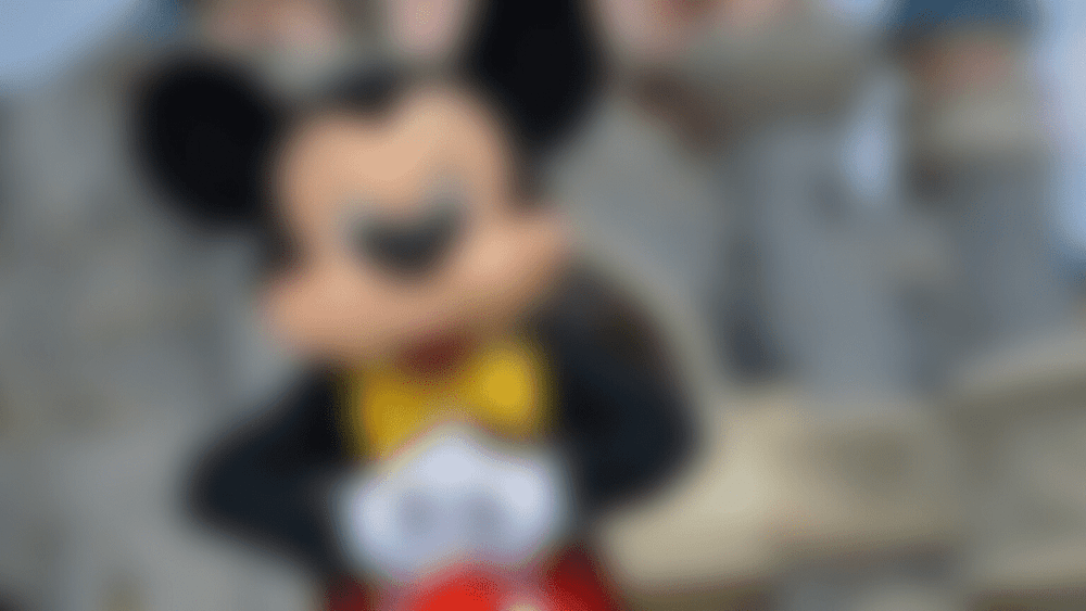 Une personne en costume de Mickey Mouse se tient devant un groupe flou de personnes à Disneyland Paris, se concentrant sur la caméra avec les grandes oreilles emblématiques et le costume rouge et noir visibles.