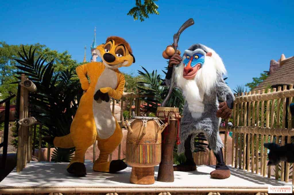 Deux personnages animés, Timon et Rafiki du Roi Lion, se tiennent à l'extérieur dans un parc à thème, Rafiki tenant un bâton et Timon posant de manière ludique près de la batterie.