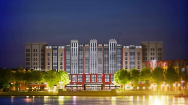 Vue nocturne d'un immeuble d'appartements moderne avec des fenêtres éclairées, comportant une section centrale rouge, flanquée d'ailes plus hautes et symétriques, se reflétant dans un plan d'eau calme au premier plan.