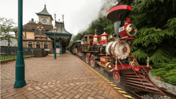 Un train à vapeur vintage et coloré du Disneyland Railroad, orné de rouge et d'or, s'est arrêté dans une gare à l'architecture victorienne ornée, sous un ciel nuageux.