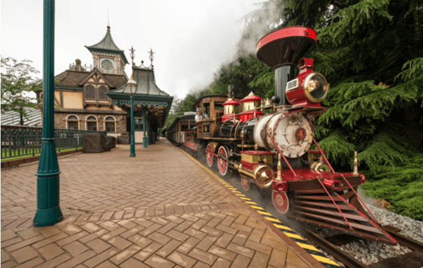 Un train à vapeur vintage et coloré du Disneyland Railroad, orné de rouge et d'or, s'est arrêté dans une gare à l'architecture victorienne ornée, sous un ciel nuageux.