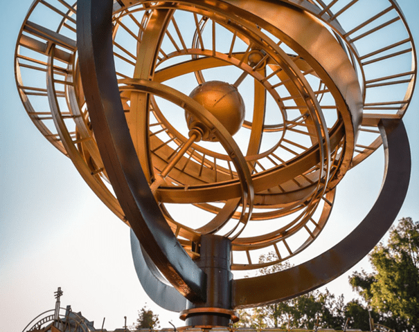 Grande sculpture cinétique comportant des anneaux métalliques circulaires et un orbe central doré, sur un ciel bleu. Le mot « Discoveryland » est affiché bien en évidence en bas.