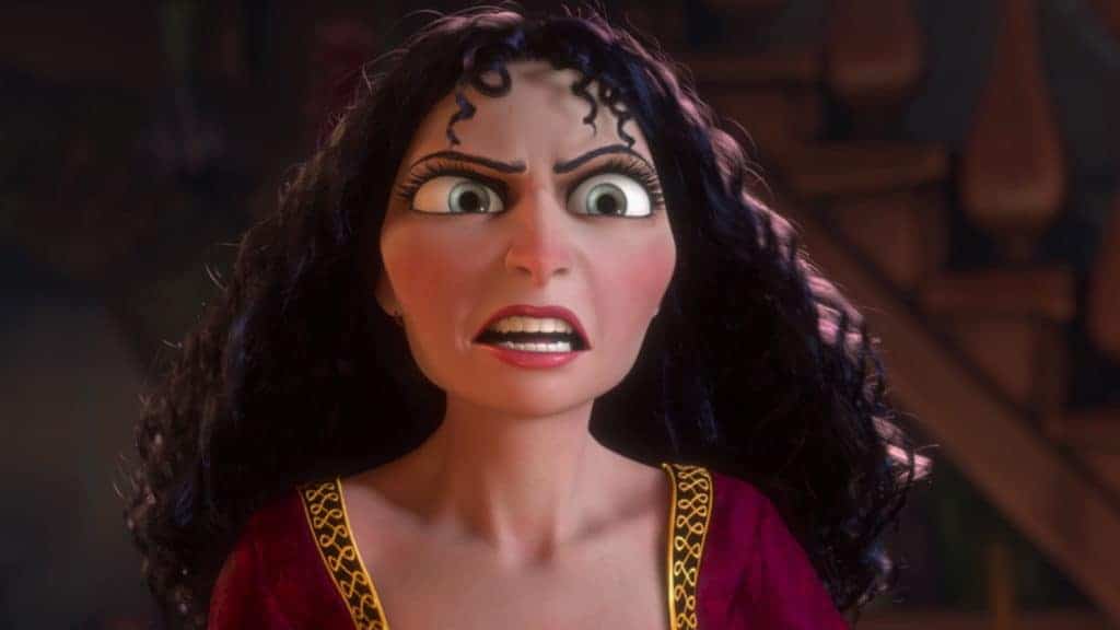 Un personnage animé aux cheveux noirs bouclés, aux yeux intenses et aux marques décoratives sur le front semble surpris dans une pièce faiblement éclairée remplie d'étagères. Elle porte une tenue rouge et or digne de Disney