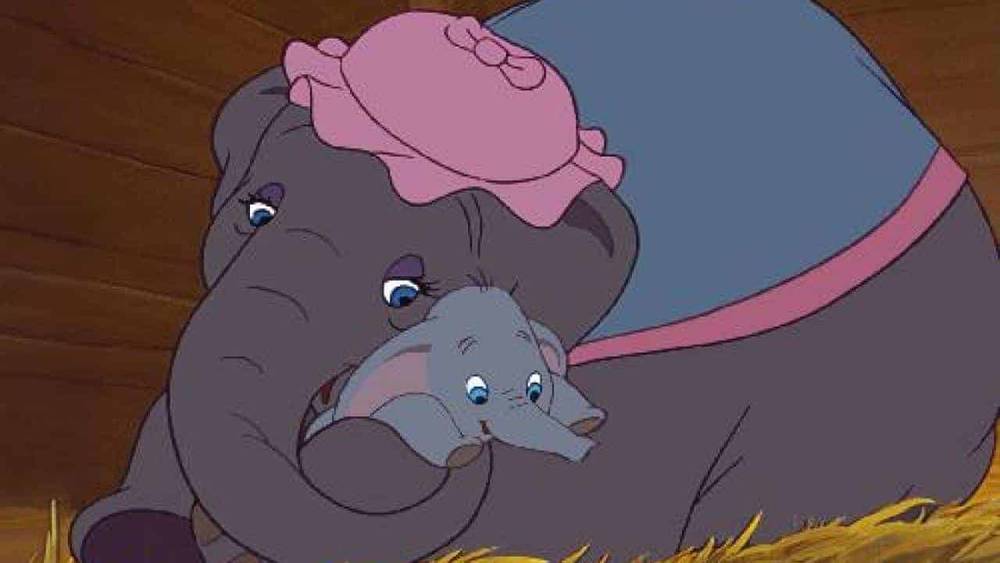 Une image tirée du film d'animation "Dumbo" montrant un bébé éléphant bercé de manière protectrice sous la trompe d'un éléphant plus grand, tous deux portant des accessoires de cirque roses.