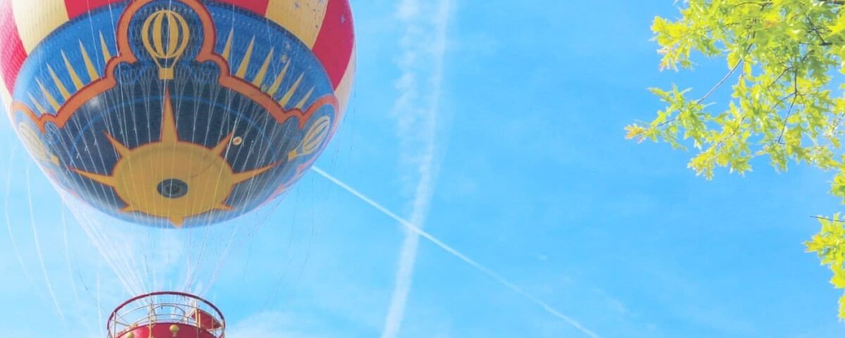 Une montgolfière colorée avec une nacelle rouge flotte près du PanoraMagique à Disneyland Paris, sous un ciel bleu à proximité de branches d'arbres vert vif.