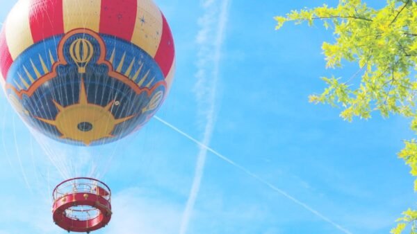Une montgolfière colorée avec une nacelle rouge flotte près du PanoraMagique à Disneyland Paris, sous un ciel bleu à proximité de branches d'arbres vert vif.