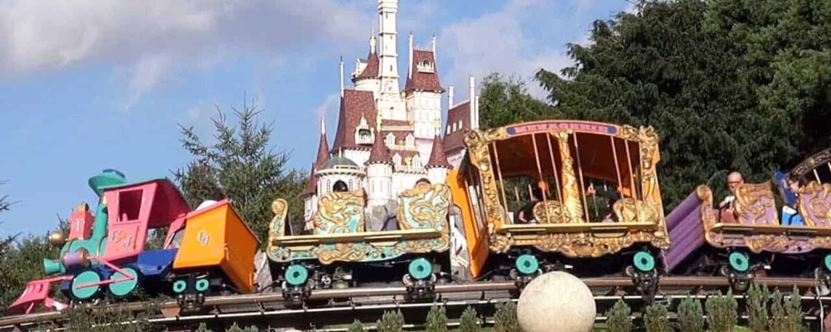 Montagnes russes colorées du parc d'attractions avec des voitures au design fantaisiste au premier plan et un château de conte de fées en toile de fond sous un ciel bleu avec des nuages duveteux ressemblant à Dumbo.