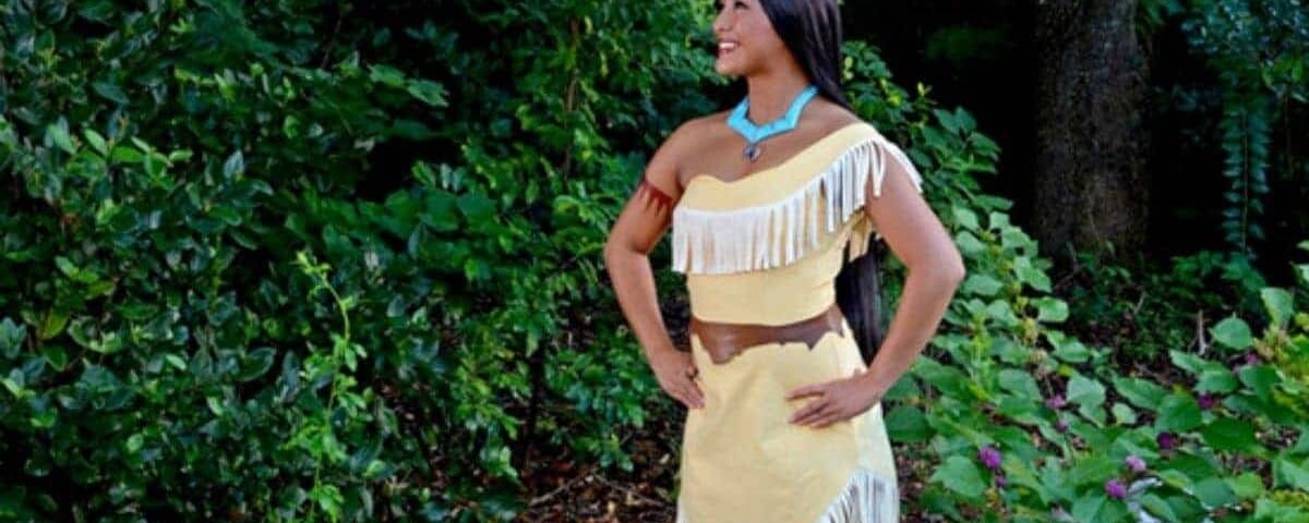 Une femme vêtue d’une tenue traditionnelle amérindienne, rappelant Pocahontas, se tient dehors avec une expression joyeuse, entourée d’un feuillage vert luxuriant. Elle porte une robe à franges et un collier turquoise