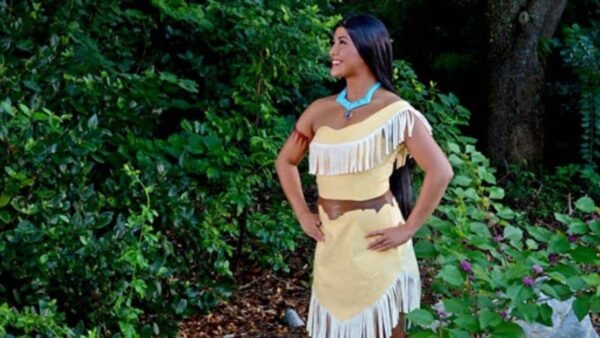 Une femme vêtue d’une tenue traditionnelle amérindienne, rappelant Pocahontas, se tient dehors avec une expression joyeuse, entourée d’un feuillage vert luxuriant. Elle porte une robe à franges et un collier turquoise