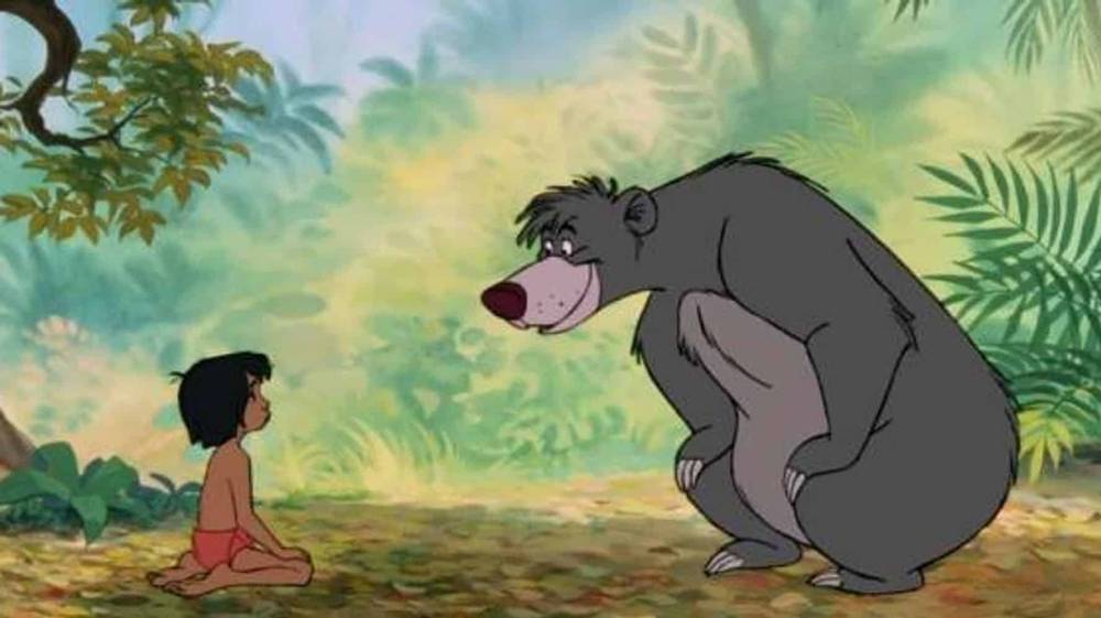 Une scène du "Livre de la Jungle" mettant en scène un jeune garçon assis en face d'un gros ours gris dans un décor de jungle luxuriante. Ils semblent converser ou partager un moment ensemble.