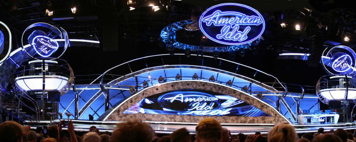 Vue panoramique de la scène de l'American Idol Experience avec des enseignes au néon bleu vif, des moniteurs circulaires affichant les candidats et un public enthousiaste.