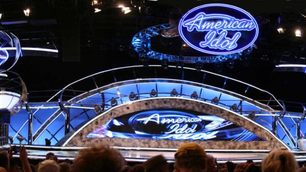 Vue panoramique de la scène de l'American Idol Experience avec des enseignes au néon bleu vif, des moniteurs circulaires affichant les candidats et un public enthousiaste.