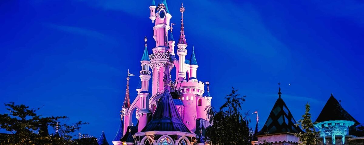 Une vue nocturne d'un château de conte de fées illuminé de lumières roses et violettes sous un ciel bleu profond, parfaite pour un séjour Disney pas cher, avec un environnement faiblement éclairé et des sentiers invitant à l'exploration.