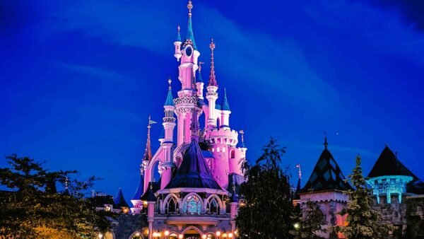Une vue nocturne d'un château de conte de fées illuminé de lumières roses et violettes sous un ciel bleu profond, parfaite pour un séjour Disney pas cher, avec un environnement faiblement éclairé et des sentiers invitant à l'exploration.