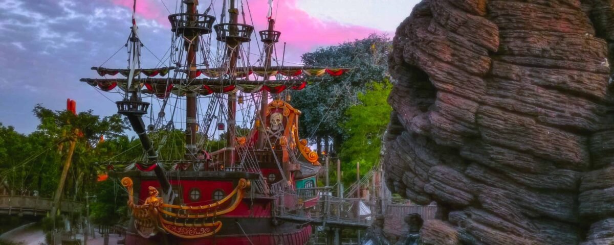 Une scène vibrante mettant en scène un grand bateau pirate aux décorations élaborées amarré à côté d'une falaise rocheuse avec de petites cascades, sous un ciel de coucher de soleil coloré à Disney.