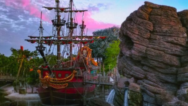 Une scène vibrante mettant en scène un grand bateau pirate aux décorations élaborées amarré à côté d'une falaise rocheuse avec de petites cascades, sous un ciel de coucher de soleil coloré à Disney.