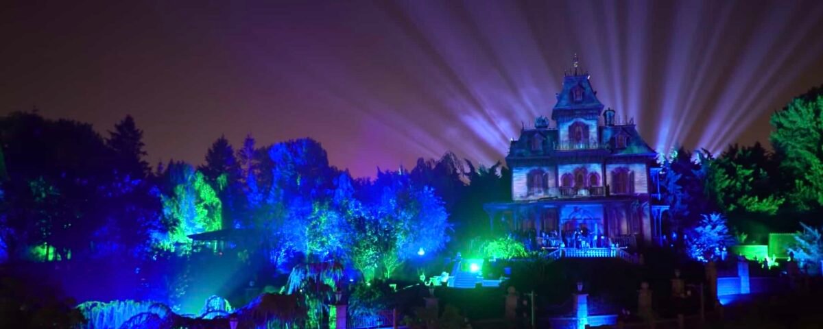 Une scène nocturne d'un grand Phantom Manor illuminé avec des faisceaux de lumière rayonnant dans le ciel, entouré de lumières vibrantes et colorées dans un jardin luxuriant à Disneyland Paris.