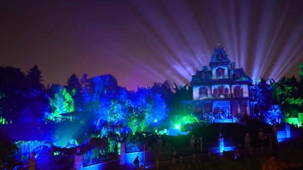 Une scène nocturne d'un grand Phantom Manor illuminé avec des faisceaux de lumière rayonnant dans le ciel, entouré de lumières vibrantes et colorées dans un jardin luxuriant à Disneyland Paris.