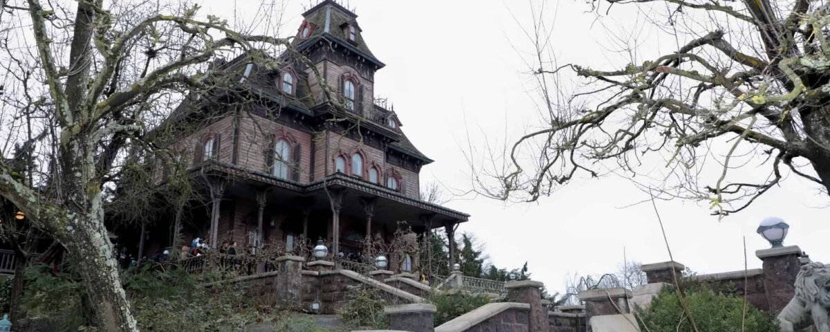 Un manoir victorien aux ornements envoûtants, inspiré du Phantom Manor de Disney, se trouve au sommet d'une colline, entouré d'arbres dénudés sous un ciel nuageux, dégageant une ambiance mystérieuse et inquiétante. Les visiteurs sont visibles
