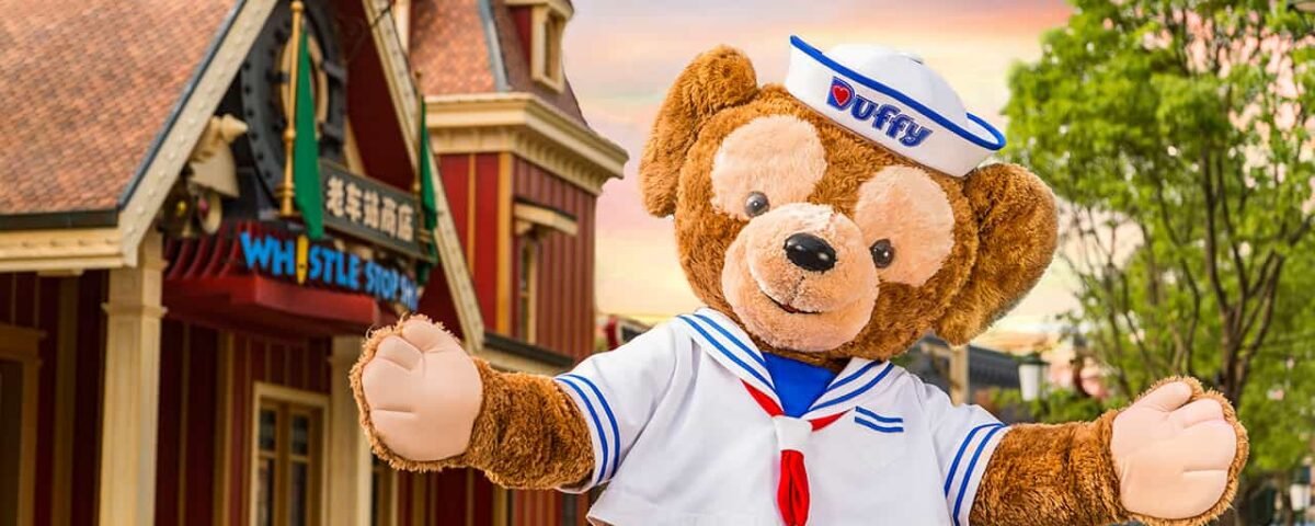 Un personnage en peluche de Duffy l'ours Disney, vêtu d'une tenue de marin avec une casquette blanche, posant joyeusement les bras tendus dans un décor de rue du parc Disneyland.