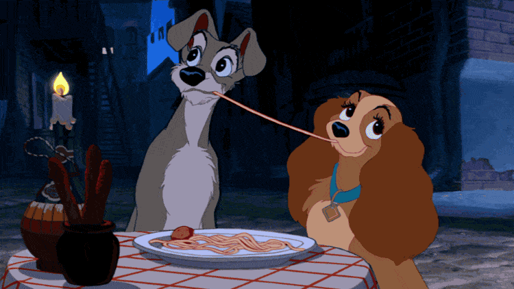 Lady et Clochard partagent des spaghettis lors d'un dîner romantique avec une bougie en fond, tirant tous deux sur le même brin, dans une scène du film romantique "La Belle et le Clochard".