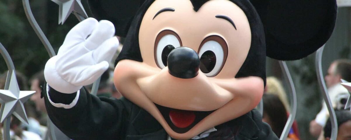 Mickey Mouse agitant joyeusement tout en portant un smoking avec un nœud papillon dans un décor de défilé Disney, avec des décorations en forme d'étoile en arrière-plan.