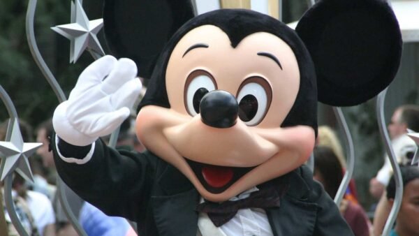 Mickey Mouse agitant joyeusement tout en portant un smoking avec un nœud papillon dans un décor de défilé Disney, avec des décorations en forme d'étoile en arrière-plan.