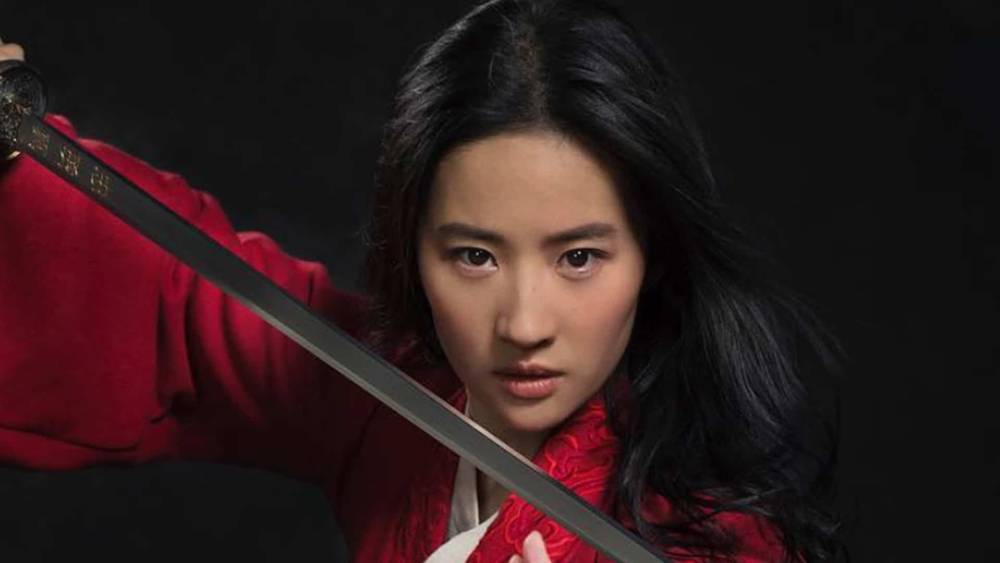 Une femme en tenue rouge, rappelant Mulan, tenant une épée pointée vers la caméra avec une expression intense. Elle a de longs cheveux noirs flottants et un air déterminé.