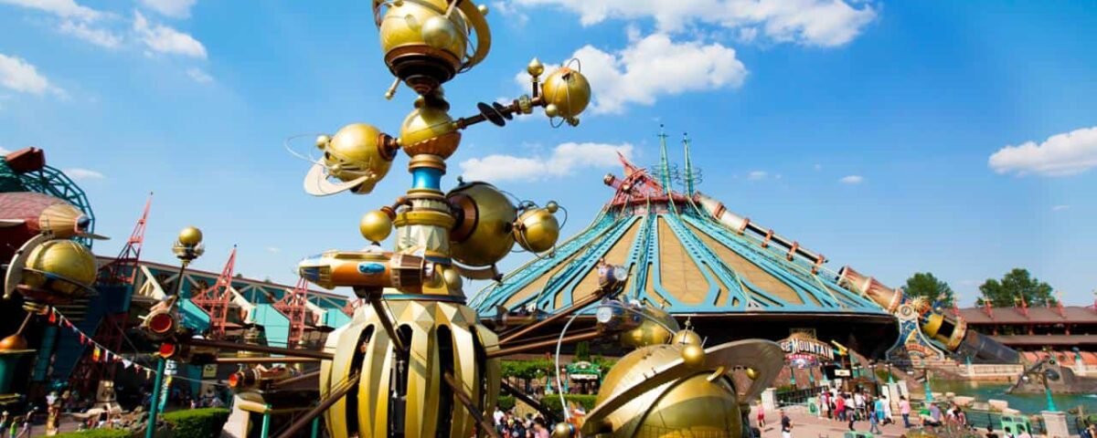 Image vibrante d'un carrousel fantaisiste d'inspiration steampunk à Disneyland Paris avec une grande foule et un bâtiment futuriste au toit pointu en arrière-plan sous un ciel bleu clair.