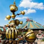 Image vibrante d'un carrousel fantaisiste d'inspiration steampunk à Disneyland Paris avec une grande foule et un bâtiment futuriste au toit pointu en arrière-plan sous un ciel bleu clair.