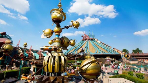 Image vibrante d'un carrousel fantaisiste d'inspiration steampunk à Disneyland Paris avec une grande foule et un bâtiment futuriste au toit pointu en arrière-plan sous un ciel bleu clair.