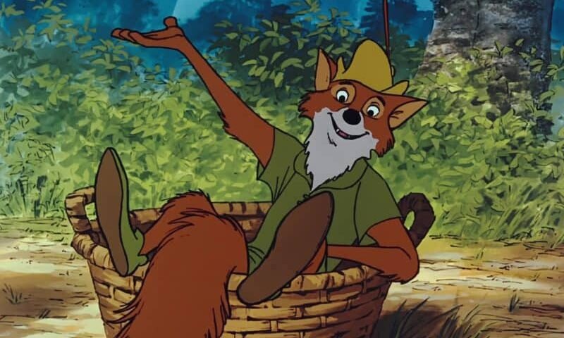 Renard animé portant une chemise verte et un chapeau jaune, allongé dans un panier dans un décor de forêt Disney, faisant des gestes avec un bras levé.