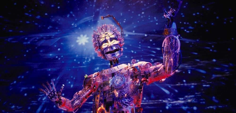 Un robot fantaisiste avec une tête en forme de dôme transparent, des fils colorés pour les cheveux et des parties mécaniques du corps, sur fond d'espace étoilé à Discoveryland, s'étendant comme s'il interagissait avec les étoiles.
