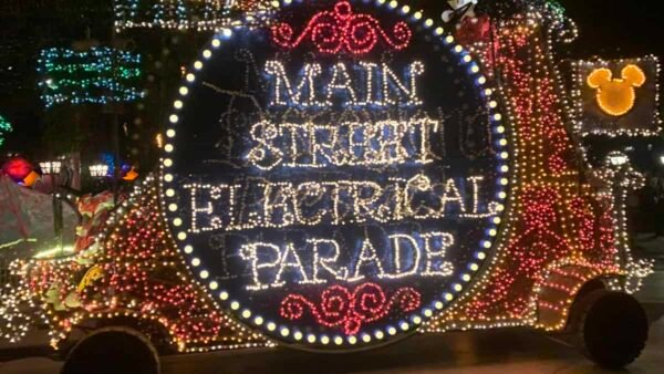 Une scène nocturne vibrante d'un char électrique de la rue principale de Disneyland, orné de lumières multicolores comportant des mots et des dessins, avec une figurine de Mickey Mouse agitant du haut.