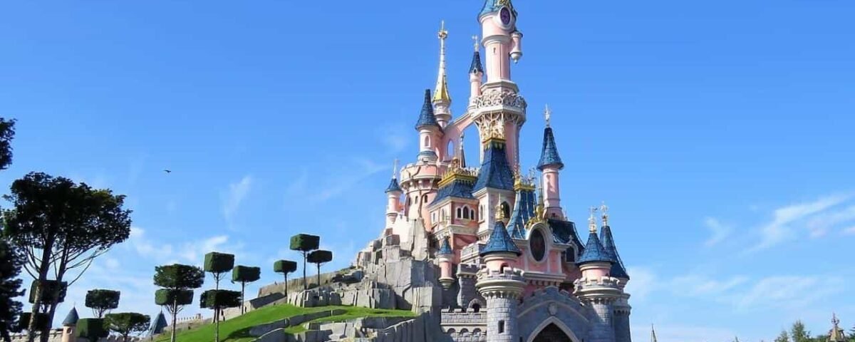 Une image vibrante du château de la Belle au Bois Dormant à Disneyland Paris, avec des flèches roses et bleues sous un ciel bleu clair avec de la verdure entourant la base.